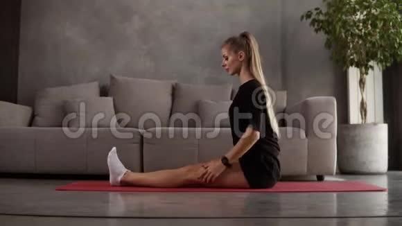 白种人金发碧眼的女人穿着黑色运动服在家里训练在灰色沙发前垫着垫子伸展着肌肉视频的预览图