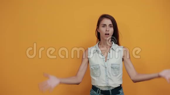 穿着蓝色牛仔衬衫的白人年轻女子震惊了双手伸向橙色的墙壁视频的预览图