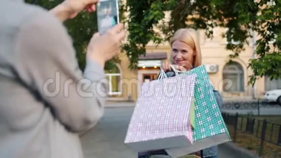 漂亮的金发美女摆着智能手机摄像头手里拿着购物袋视频的预览图