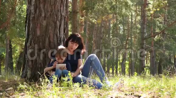 母亲和儿子花时间坐在森林里的一棵树旁女人和孩子在平板电脑里玩视频的预览图