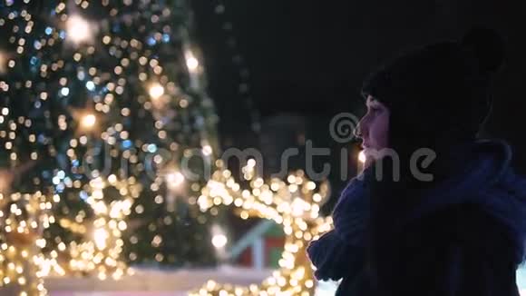 年轻迷人的女人站在闪亮的圣诞树前穿着温暖衣服的女人在外面看着夜晚视频的预览图