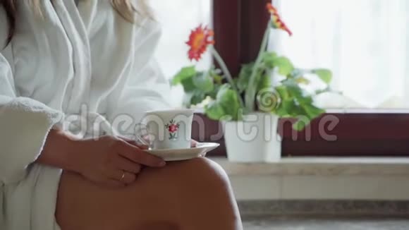 特写身穿白色bahrobe的女人坐在窗边腿上端着一杯白色的茶或咖啡咖啡休息时间视频的预览图