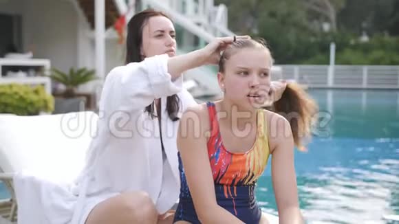 穿着白色浴袍的女人梳着女儿头发坐在别墅游泳池旁的躺椅上视频的预览图