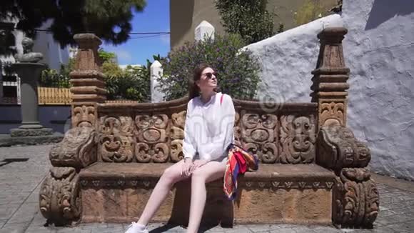 美丽的女游客戴着眼镜坐在一张古老的雕刻长凳上阳光明媚的一天街上的石凳视频的预览图