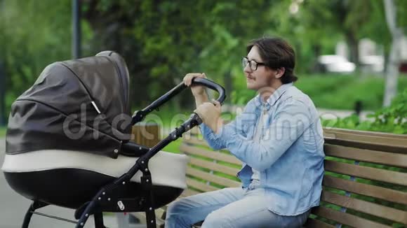 快乐的年轻爸爸欣赏刚出生的摇摆婴儿车坐在公园的长凳上视频的预览图