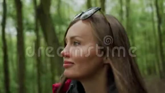 美丽的微笑女人穿着红色夹克背包在茂密的森林里散步环顾四周享受大自然的美丽视频的预览图