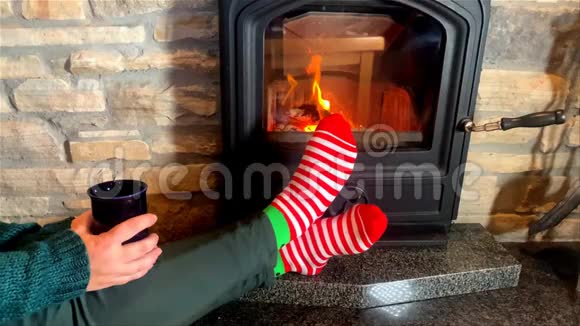 有一个杯子的女人坐在滚烫的壁炉旁双腿穿着红色条纹袜子伸到炉火旁冬季放松的概念视频的预览图