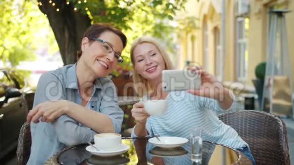 金发美女与朋友在户外咖啡厅自拍用智能手机端着咖啡视频的预览图