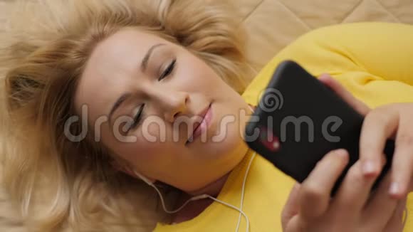 一个美丽的女人躺在床上一边用智能手机听音乐视频的预览图