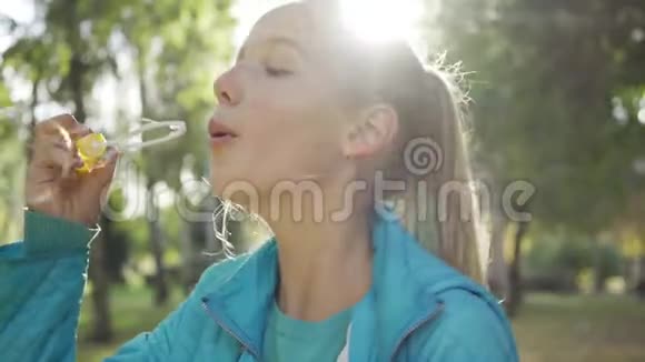 在阳光明媚的公园里微笑的白种人青少年吹着肥皂泡穿着休闲装的金发少女视频的预览图