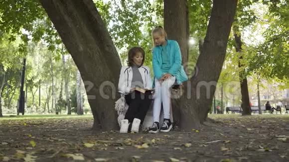 美丽的黑发白种人女人读这本书给一个年轻的金发女孩在阳光明媚的公园两个穿休闲服装的人视频的预览图