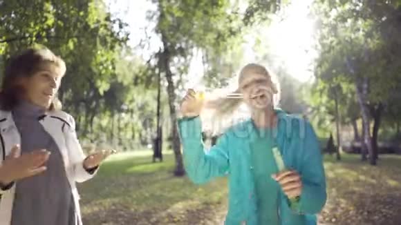 快乐的少女白种人女孩吹肥皂泡给她积极的祖母在阳光明媚的秋天公园家庭成员视频的预览图