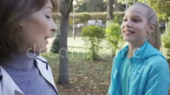 一个年轻的微笑金发女孩的特写听着这位年长的黑发女人一家人在秋天公园散步视频的预览图
