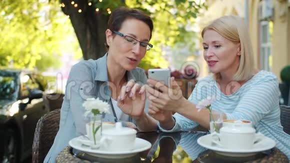 快乐的女士们笑着在户外咖啡馆里看着智能手机屏幕视频的预览图