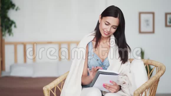 年轻女子裹着格子坐在椅子上在笔记本上做笔记视频的预览图