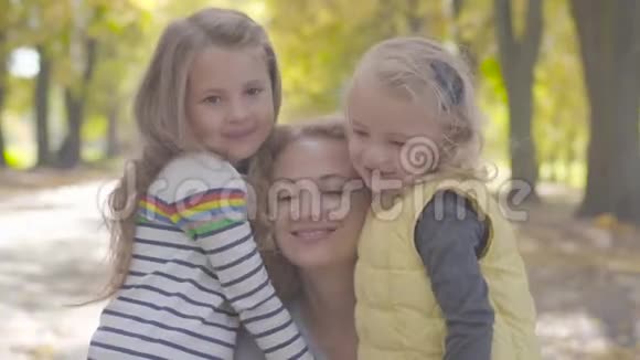在秋天公园里两个快乐的白种人小妹妹拥抱着卷曲的金发女人的特写肖像可爱的微笑视频的预览图