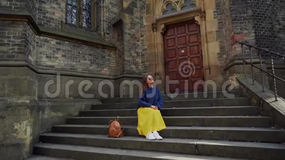 美丽的女孩穿着一条黄色长裙背着背包坐在靠近天主教会的台阶上总计划视频的预览图
