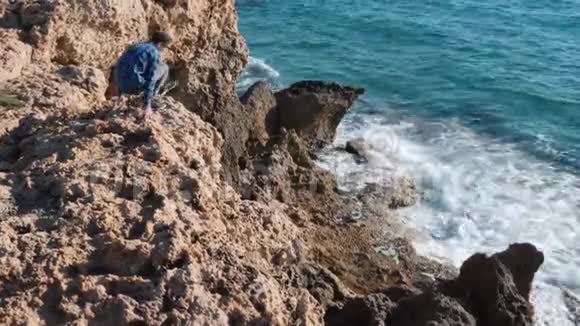 可爱的白种人坐在悬崖边上的岩石上小男孩走下悬崖望着大海他坐在岩石海岸视频的预览图