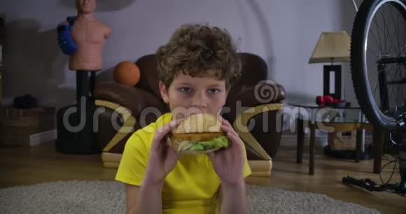 白人男孩看电视吃汉堡包的肖像可爱的卷发少年家里有垃圾食品休闲视频的预览图