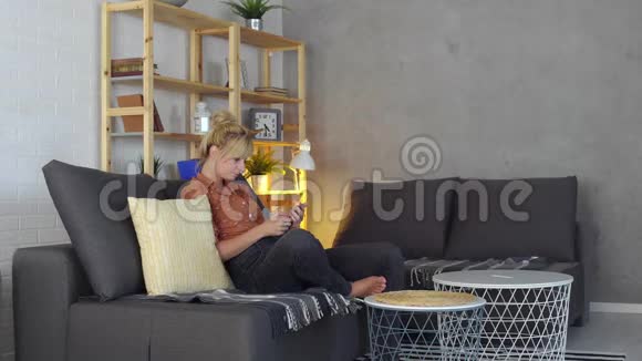 一位年轻的金发休闲女士正在用她的电话在客厅里度过一些空闲时间视频的预览图
