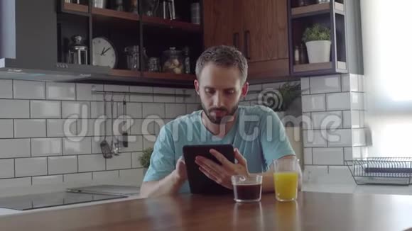 有魅力的帅哥留着胡子在厨房的平板电脑上看比赛成绩视频的预览图