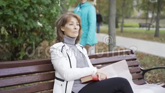 一个年轻的金发女孩来到阳光明媚的公园里坐在长凳上的老妇人身边年轻漂亮的黑发女人视频的预览图