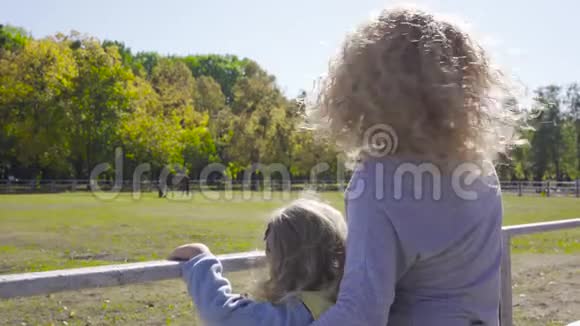 漂亮的金发卷发女人和两个小女孩站在畜栏旁边妈妈给她可爱的马送马视频的预览图