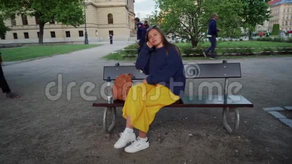 穿着黄色长裙的漂亮女孩坐在公园的长凳上有时她会弯下眼睛视频的预览图