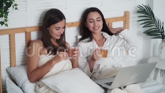 两个女朋友坐在床上喝茶在笔记本电脑上看电影聊天视频的预览图