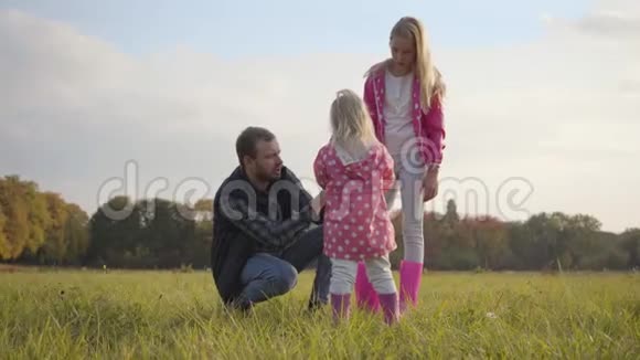 年轻的白种人在秋天的晚上在户外和他的女儿们交谈父亲和两个穿粉色衣服的金发女孩视频的预览图