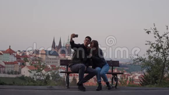 可爱的情侣在电话上自拍坐在长凳上美丽的布拉格景色视频的预览图