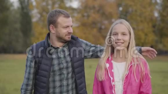 微笑的白种人男人走近前景上的年轻金发女孩拥抱她父亲在做一件视频的预览图