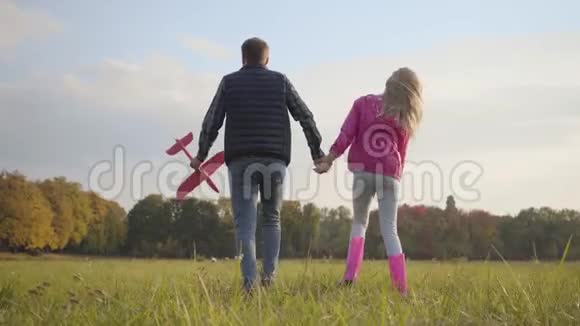 一个白人男子牵着一个年轻的金发女孩和粉红色玩具飞机的手他父亲带着他可爱的孩子视频的预览图