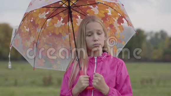 悲伤的白种人女孩一头金发一双浅棕色的眼睛穿着粉红色的夹克拿着一把彩色的伞视频的预览图