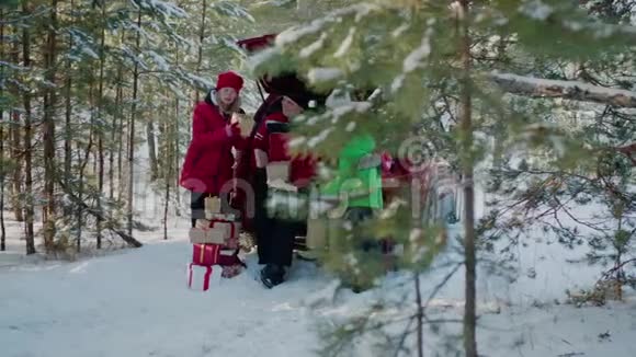 在冬季森林的汽车后备箱里寻找新年礼物的家庭快乐妈妈爸爸儿子和女儿戴着红色圣诞帽视频的预览图