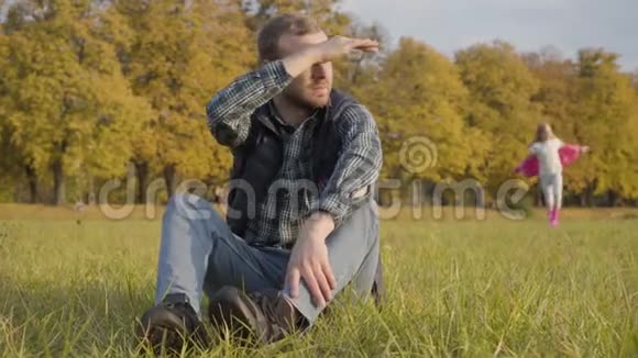 微笑的白种人女孩跑到一个男人坐在秋天的草地上穿着休闲服装的金发少女视频的预览图