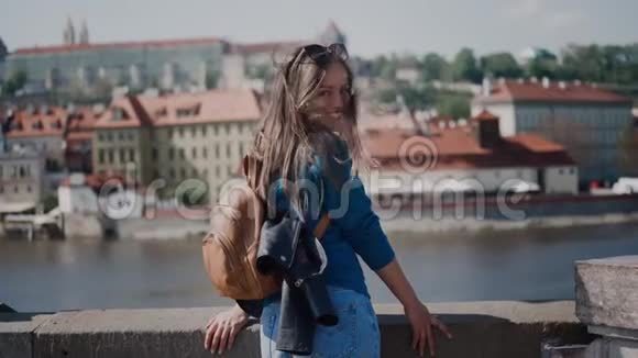 背着背包的女孩站在桥上微笑着看着摄像机在河流的背景下在古老的城市中风景如画视频的预览图