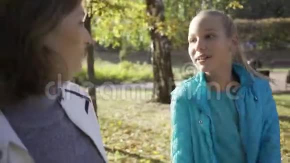 年轻的微笑金发女孩与年长的黑发女人交谈家人在秋天公园散步聊天奶奶视频的预览图