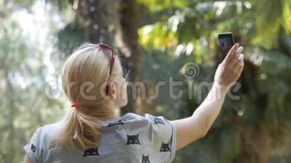 一位穿过热带公园的妇女使用智能手机相机拍摄棕榈树和其他植物视频的预览图