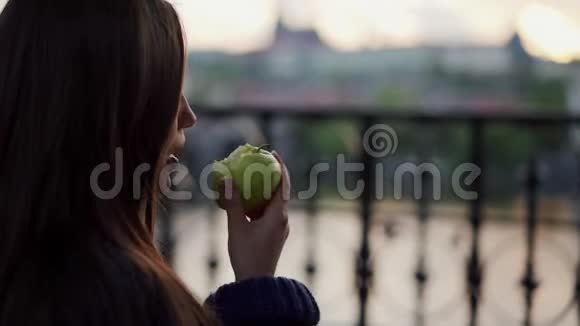 留着长发的布鲁内特在城市风景模糊的背景下咬着绿苹果视频的预览图
