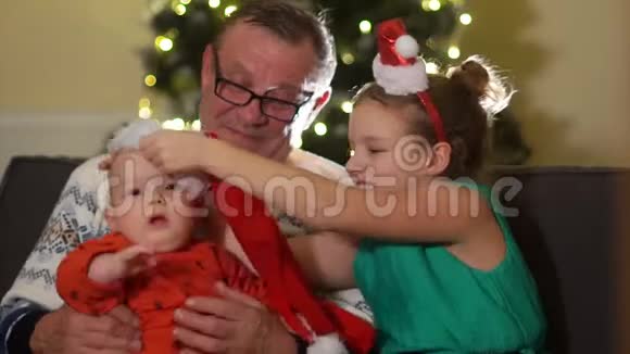 祖父带着孙子在沙发上坐圣诞姐姐把婴儿戴在圣诞老人的帽子上宝宝惊喜视频的预览图