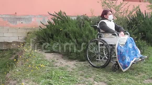 残疾妇女独自坐在轮椅上戴着保护面具在花园里向右看但被隔离视频的预览图