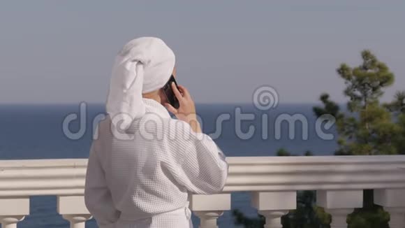 一个穿着白色长袍头上戴着毛巾的女人站在一个豪华的露台上俯瞰大海在打电话视频的预览图