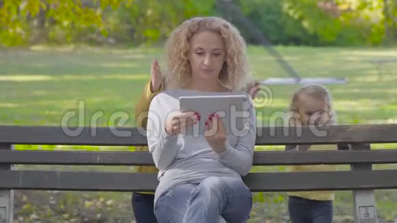年轻漂亮的白种人女人坐在长凳上拿着一块平板电脑两个小可爱的女孩从后面走过来然后关上了视频的预览图