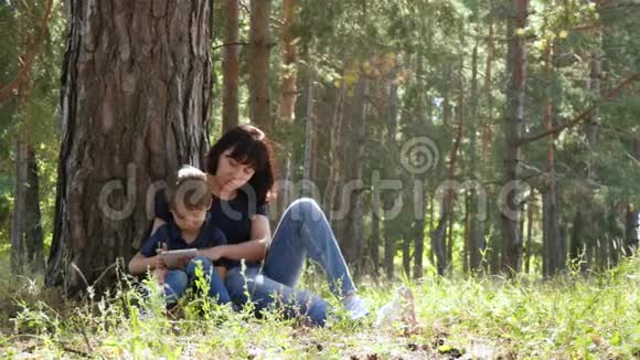 幸福家庭母子俩坐在树边的公园里放松一个女人和一个孩子看着平板电脑屏幕视频的预览图