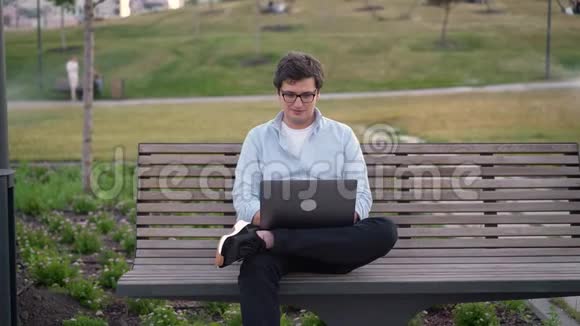 公园里坐在草地模糊的长凳上用笔记本电脑的年轻人视频的预览图