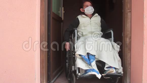 残疾妇女独自一人坐在轮椅上戴着防护面罩望着外面的敞开的房门呆在家里进行检疫视频的预览图