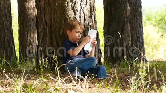 一个可爱的小男孩坐在公园的一棵树旁拿着一块平板电脑一个快乐的孩子用平板电脑打电话视频的预览图