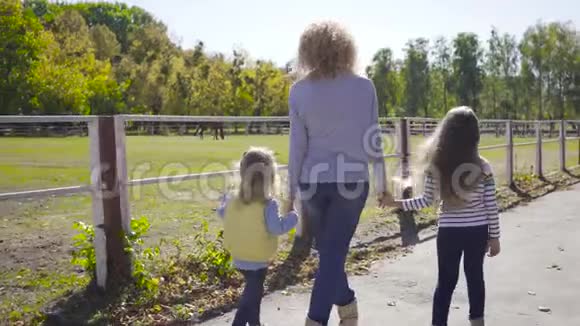 从后面可以看到金发白种人的女人和她的女儿一起走在马厩里美丽的棕马视频的预览图
