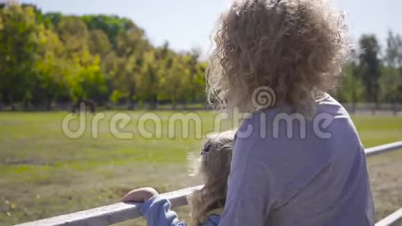 侧面看着金发卷曲的女人和小白种人的女孩站在畜栏旁边妈妈给她看马视频的预览图